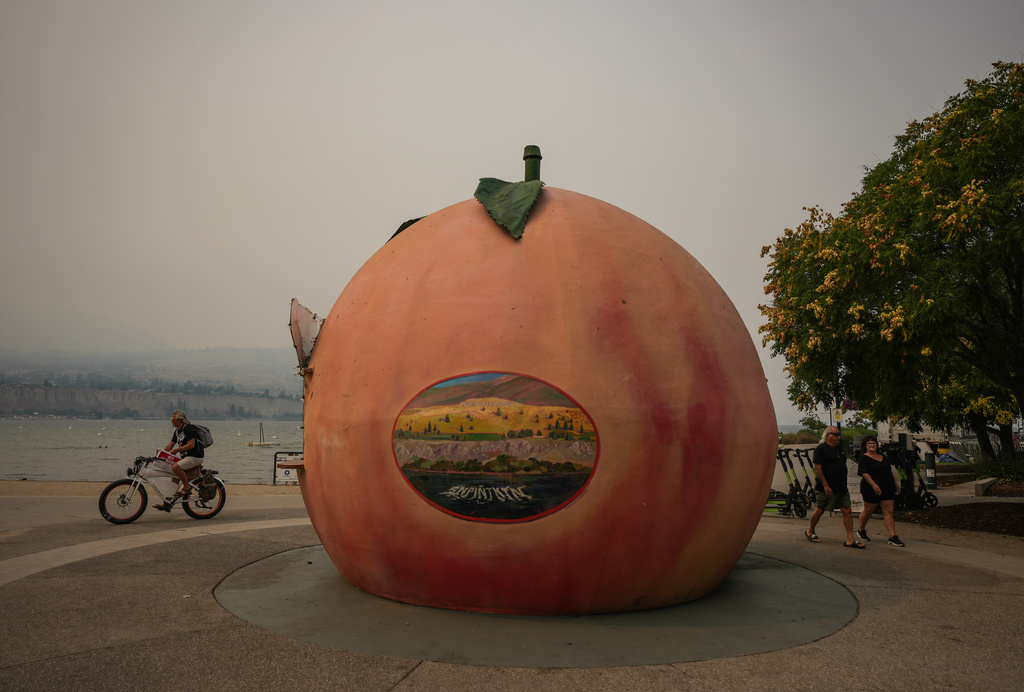 People walk and cycle past an ice cream stand in the shape of a peach as smoke from the Bald Range wildfire envelops Okanagan Lake, in Penticton, British Columbia, on Sunday, Aug. 9, 2026. (Darryl Dyck/The Canadian Press via AP)