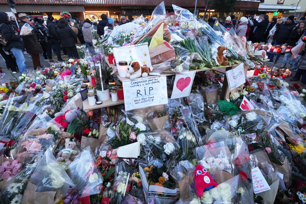 People stand around floral tributes and candles placed outside the sealed off Le Constellation bar in Crans-Montana, Swiss Alps, Switzerland, Saturday, Jan. 3, 2026, where a devastating fire left dead and injured during the New Year's celebrations. (AP Photo/ Antonio Calanni)