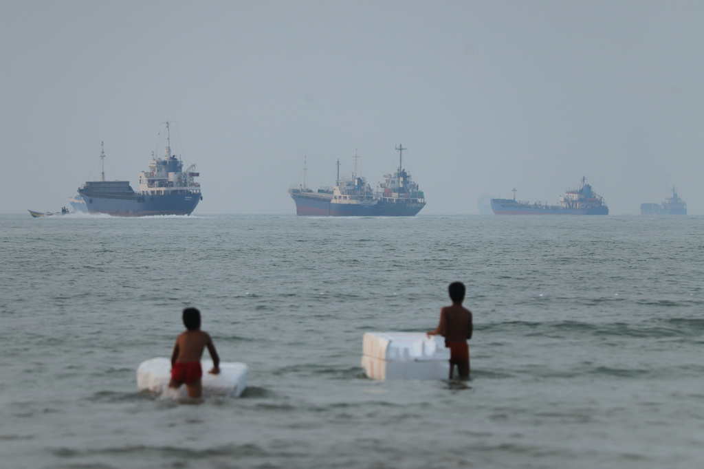 Two boys stand in shallow water with foam floats as cargo ships and other vessels are seen in the Strait of Hormuz off Bandar Abbas, Iran, Tuesday, June 30, 2026. (Amirhosein Khorgooi/ISNA via AP)