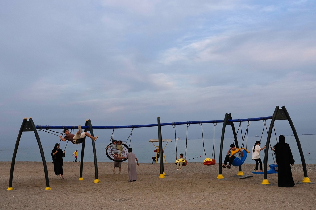 Children and adults play on swings on the beach as oil tankers and cargo ships line up in the Strait of Hormuz near Khor Fakkan, United Arab Emirates, Wednesday, March 11, 2026. (AP Photo/Altaf Qadri)
