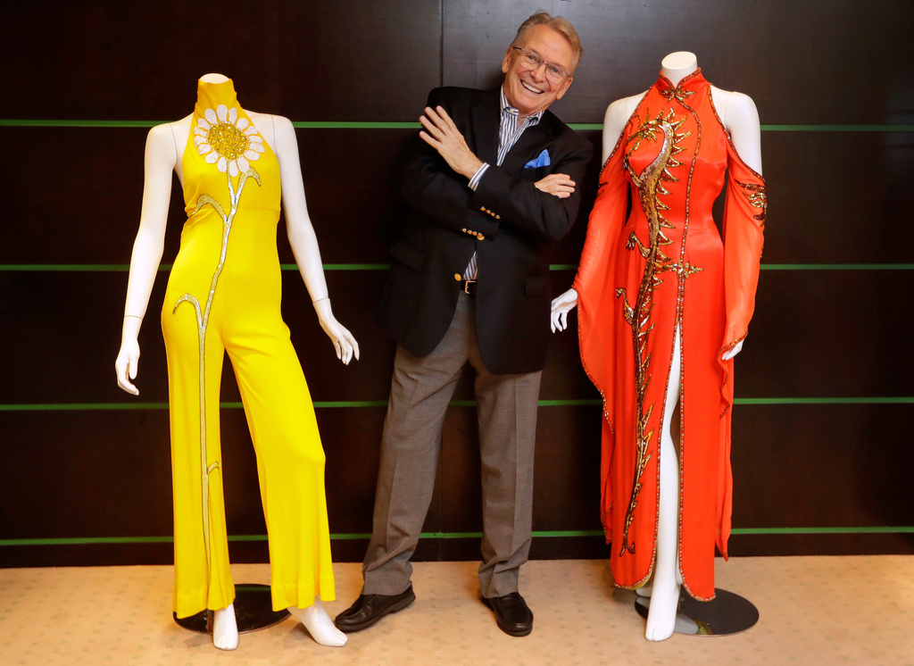 FILE - Fashion and Costume Designer Bob Mackie poses with two of his iconic designs, a scarlet red satin gown worn by Cher in 1975, right, and a marigold jersey jumpsuit worn by Cher between 1971-1976, left, in London on Aug. 16, 2018. (AP Photo/Kirsty Wigglesworth, File)