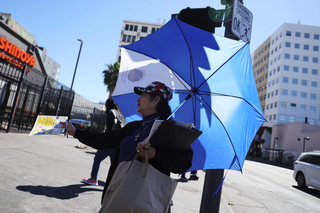 A woman covers herself from the sun while handing out fliers during an unseasonably hot day at MacArthur Park on Thursday, March 12, 2026, in Los Angeles. (AP Photo/Ryan Sun)