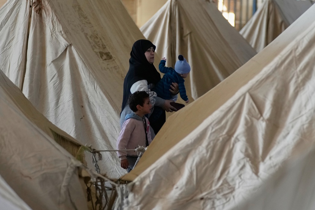A displaced woman holds a child as another stands beside her between rows of tents at the Camille Chamoun Sports City Stadium, which has been turned into a shelter for people displaced by Israeli airstrikes in southern Lebanon and Dahiyeh, Beirut's southern suburbs, in Beirut, Lebanon, Tuesday, March 10, 2026. (AP Photo/Hassan Ammar)