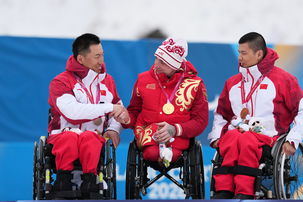 Silver medalist Mao Zhongwu, of China, left, shakes hands with gold medalist Ivan Golubkov, of Russia, while bronze medalist Zheng Peng, of China, looks on, on the podium after the cross country skiing men's 10Km interval start sitting final at the 2026 Winter Paralympics, in Tesero, Italy, Wednesday, March 11, 2026. (AP Photo/Evgeniy Maloletka)