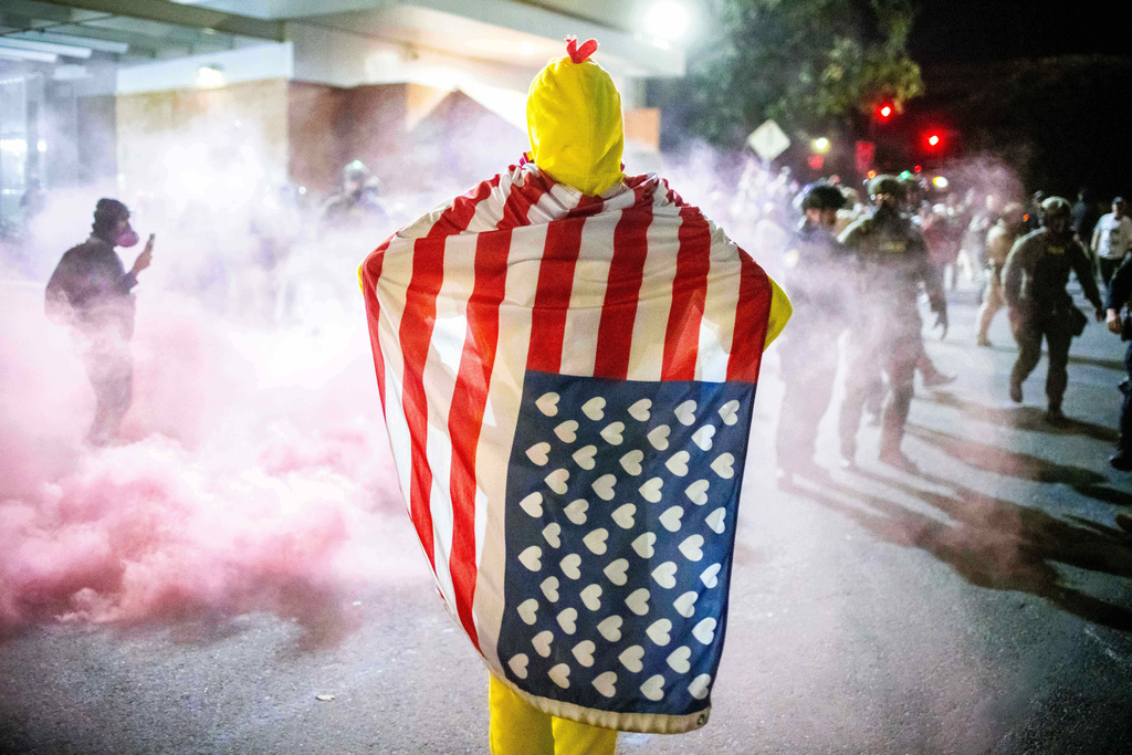 A protester stands draped in an American flag watches as law enforcement officers try to disperse protesters near a U.S. Immigration and Customs Enforcement facility in Portland, Ore. on Sunday, Oct. 5, 2025. (AP Photo/Ethan Swope)