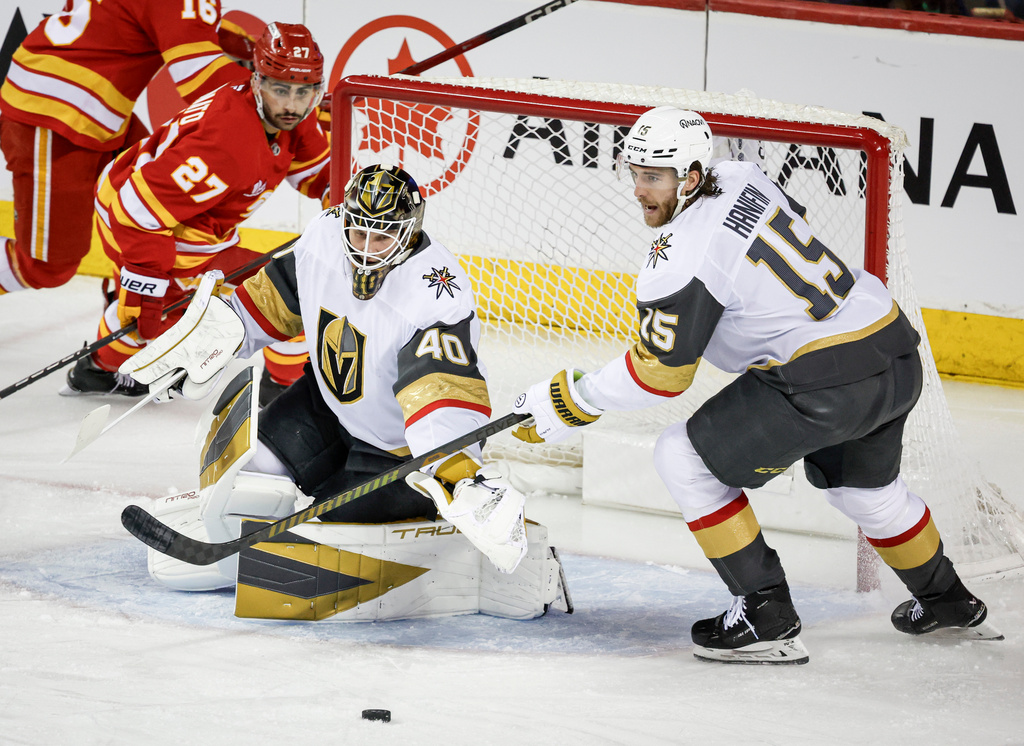 Vegas Golden Knights goalie Akira Schmid, center, and Noah Hanifin (15) reach for the puck as Calgary Flames' Matt Coronato (27) looks on during second-period NHL hockey game action in Calgary, Saturday, Dec. 20, 2025. (Jeff McIntosh/The Canadian Press via AP)