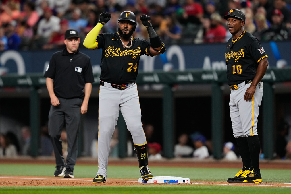 Pittsburgh Pirates' Marcell Ozuna (24) celebrates a run-scoring single as first base coach Tarrik Brock (16) and umpire Dan Merzel, left, look on in the first inning of a baseball game against the Texas Rangers Wednesday, April 22, 2026, in Arlington, Texas. (AP Photo/Tony Gutierrez)