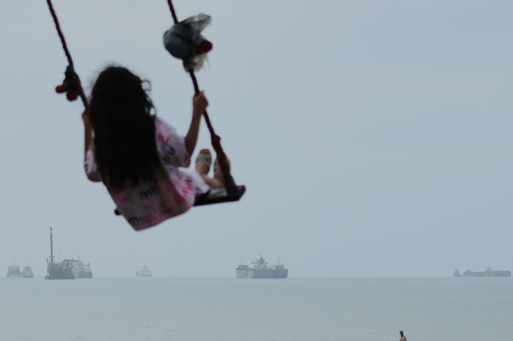A girl swings along the shore as cargo ships and other commercial vessels appear anchored in the Strait of Hormuz off Bandar Abbas, Iran, Monday, July 27, 2026. (Razieh Poudat/ISNA via AP)