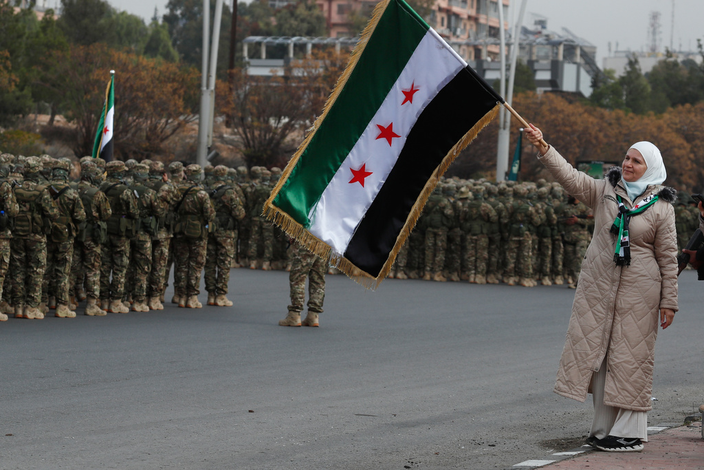 A woman waves the Syrian flag as members of the new Syrian army stand in formation during celebrations marking the first anniversary of the ousting of former President Bashar Assad in Damascus, Syria, Monday, Dec. 8, 2025. (AP Photo/Omar Sanadiki)
