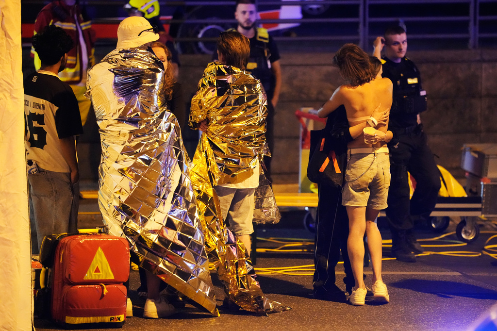 Two people embrace near others wearing thermal blankets as they stand near emergency personnel in Berlin, early Sunday, July 26, 2026, following the cancellation of Christopher Street Day festivities. (AP Photo/Markus Schreiber)