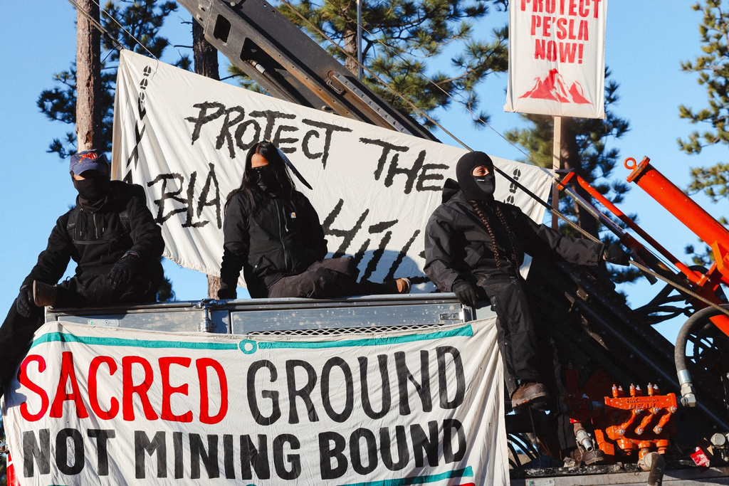 Masked demonstrators sit atop drilling equipment on Thursday, April 30, 2026, in Pennington County, S.D., with a banner reading Sacred ground not mining bound. (Angel White Eyes via AP)