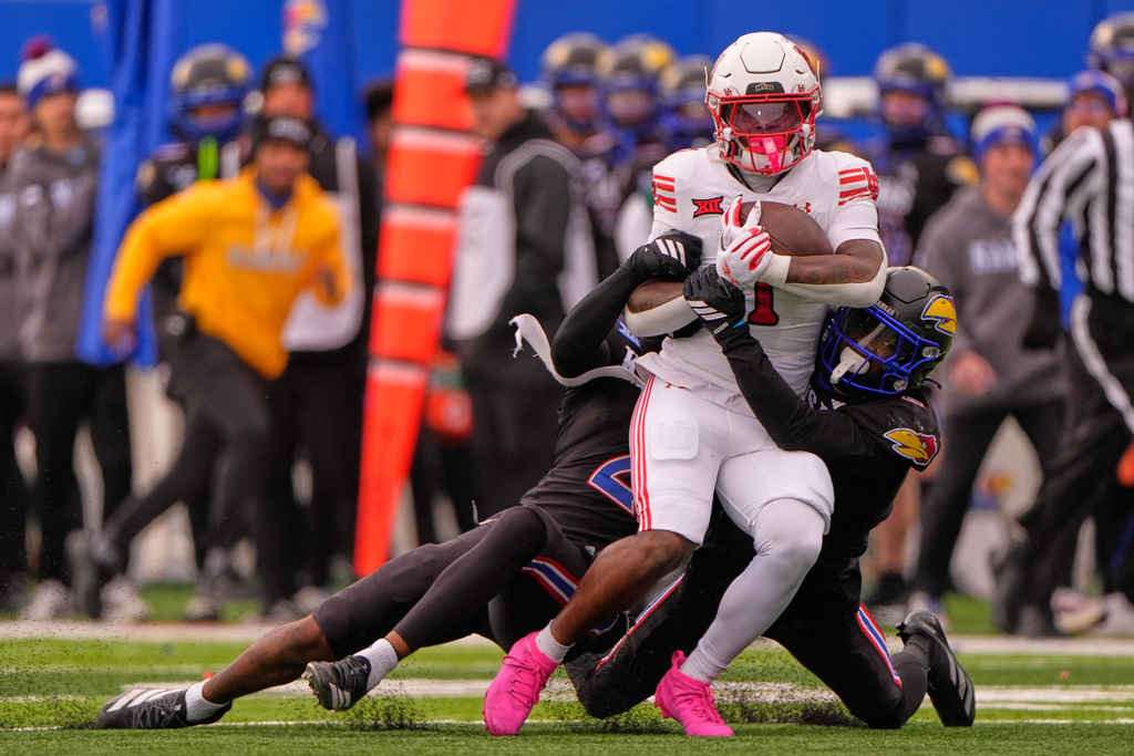 Utah running back Wayshawn Parker, center, is tackled by Kansas safety Lyrik Rawls, left, and defensive back Austin Alexander, right, during the first half of an NCAA college football game Friday, Nov. 28, 2025, in Lawrence, Kan. (AP Photo/Charlie Riedel)