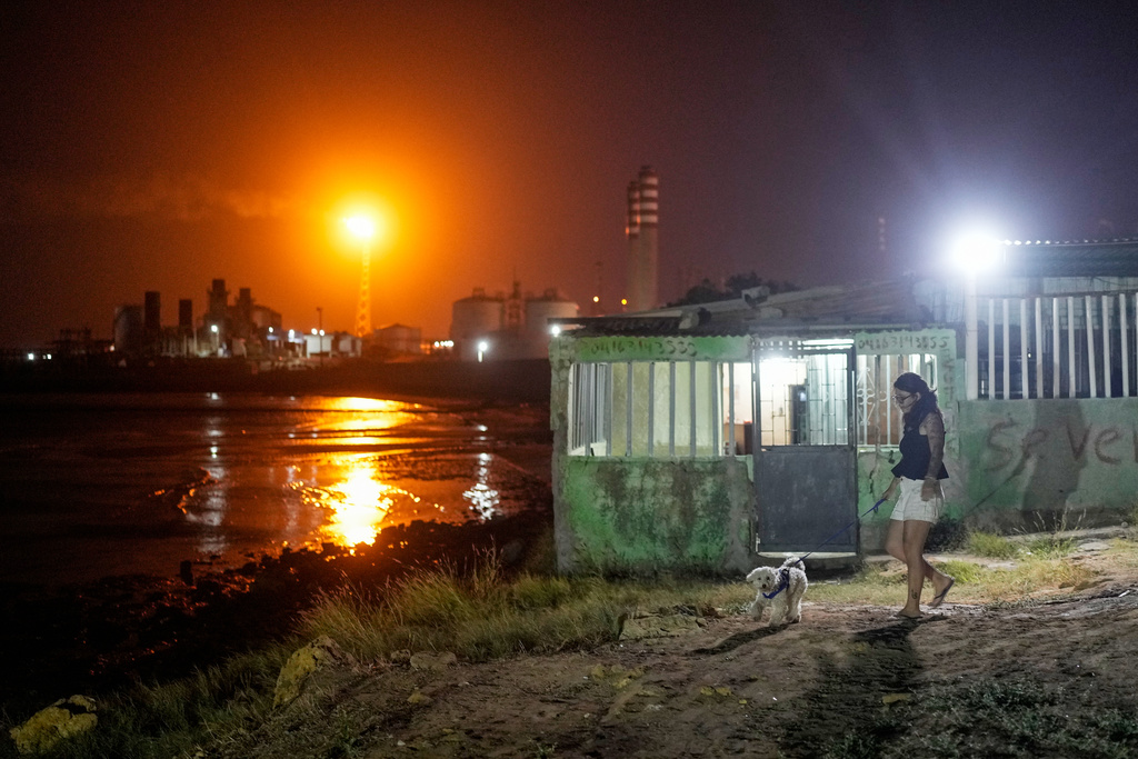 A woman walks her dog as flames burn from flare stacks at the Cardon refinery in Punta Cardon, Venezuela, Wednesday, Jan. 14, 2026. (AP Photo/Matias Delacroix)