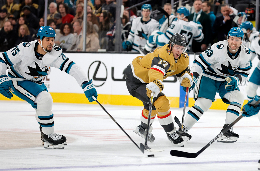 Vegas Golden Knights defenseman Ben Hutton (17) reaches for the puck between San Jose Sharks right wing Ryan Reaves (75) and center Barclay Goodrow (23) during the second period of an NHL hockey game Tuesday, Dec. 23, 2025, in Las Vegas. (AP Photo/Steve Marcus)