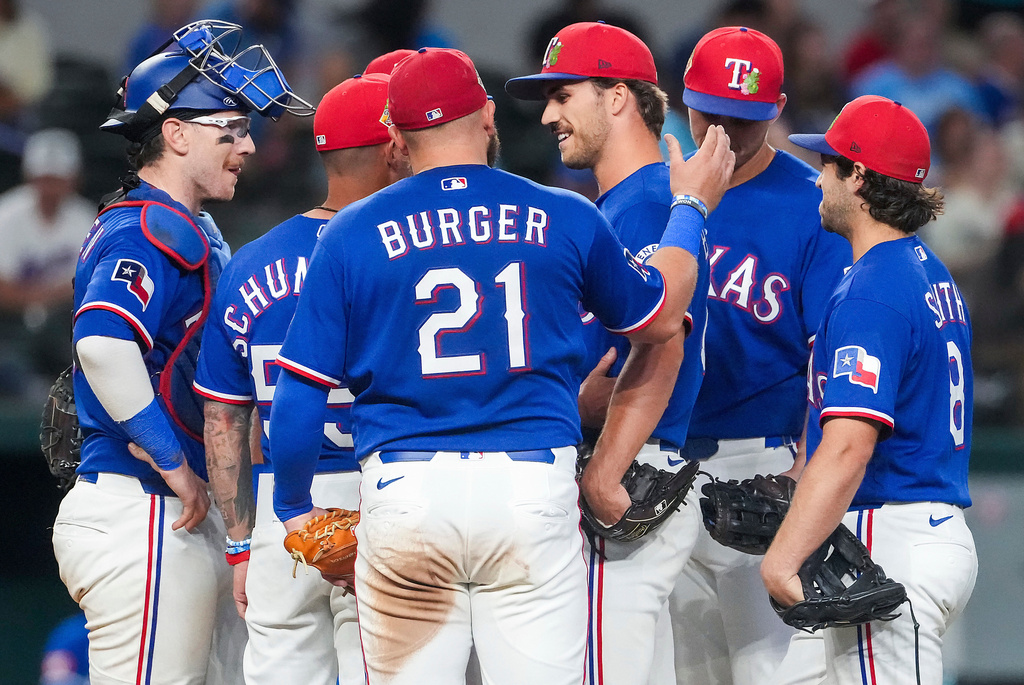 Texas Rangers pitcher Carter Baumler, third from right, gets a pat on the back from first baseman Jake Burger (21) as he gets a visit at the mound from manager Skip Schumaker, second from left, to inform him he has made the opening day roster during the fifth inning of an exhibition baseball game against the Kansas City Royals, Monday, March 23, 2026, in Arlington, Texas. (Smiley N. Pool/The Dallas Morning News via AP)