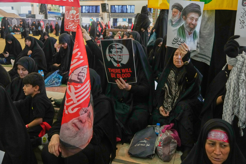 A woman holds a poster depicting U.S. President Donald Trump in the crosshairs of a rifle scope with the English words "There Will Be Blood," while another woman holds a portrait of the late Supreme Leader Ali Khamenei and his son, current Supreme Leader Mojtaba Khamenei, as mourners gather to commemorate the late leader at the Imam Khomeini Mosalla Grand Mosque in Tehran, Iran, Tuesday, July 14, 2026. (AP Photo/Vahid Salemi)
