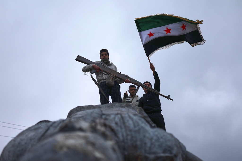 Residents wave a Syrian flag atop a toppled statue of a female Kurdish fighter, with one holding a statue's replica AK-47 that was part of the statue, after the takeover of the town by Syrian government forces from U.S.-backed Syrian Democratic Forces (SDF), in Tabqa, eastern Syria, Sunday, Jan. 18, 2026. (AP Photo/Ghaith Alsayed)
