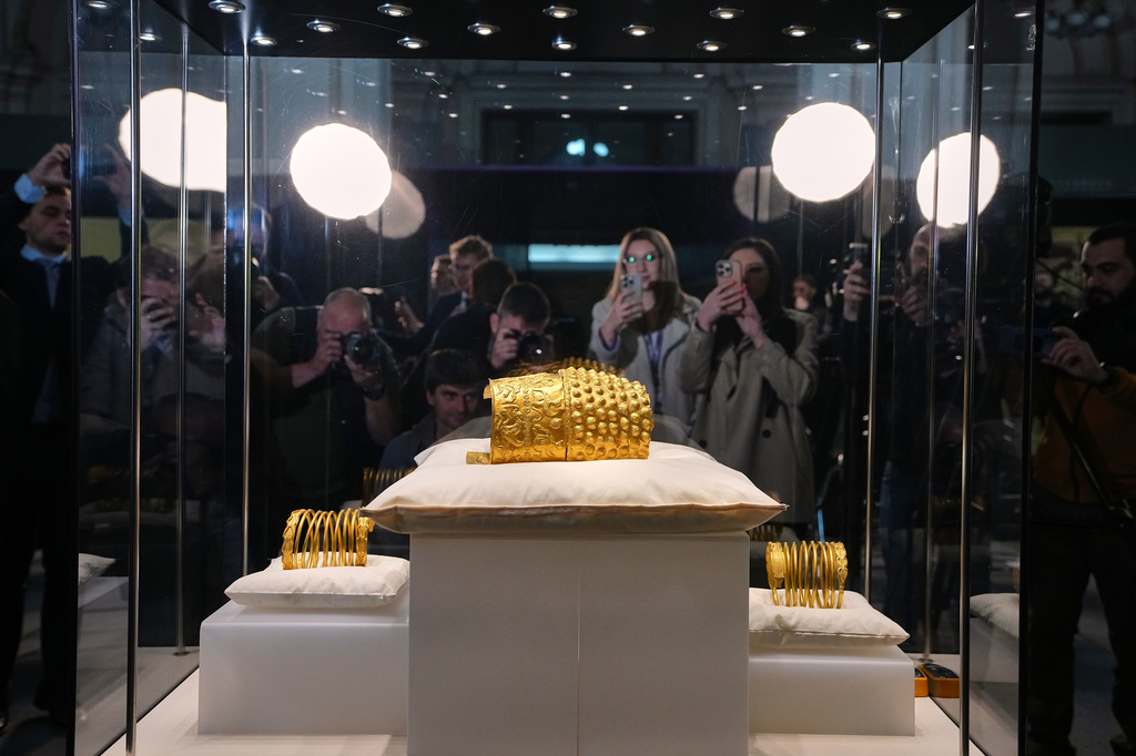 Journalists gather around a glass enclosure with the Dacian gold items, a 2,500-year-old helmet and wristbands, stolen from a museum in the Netherlands and then recovered by Dutch authorities, after they were presented upon being returned at the National Museum of Romanian History, in Bucharest, Romania, Tuesday, April 21, 2026. (AP Photo/Andreea Alexandru)