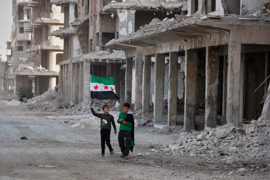 Two boys walk down a street carrying Syrian flags in the war-damaged Damascus suburb of Daraya, Syria, Friday, Dec. 5, 2025, ahead of celebrations marking the first anniversary of the ousting of the Bashar Assad regime. (AP Photo/Omar Sanadiki)