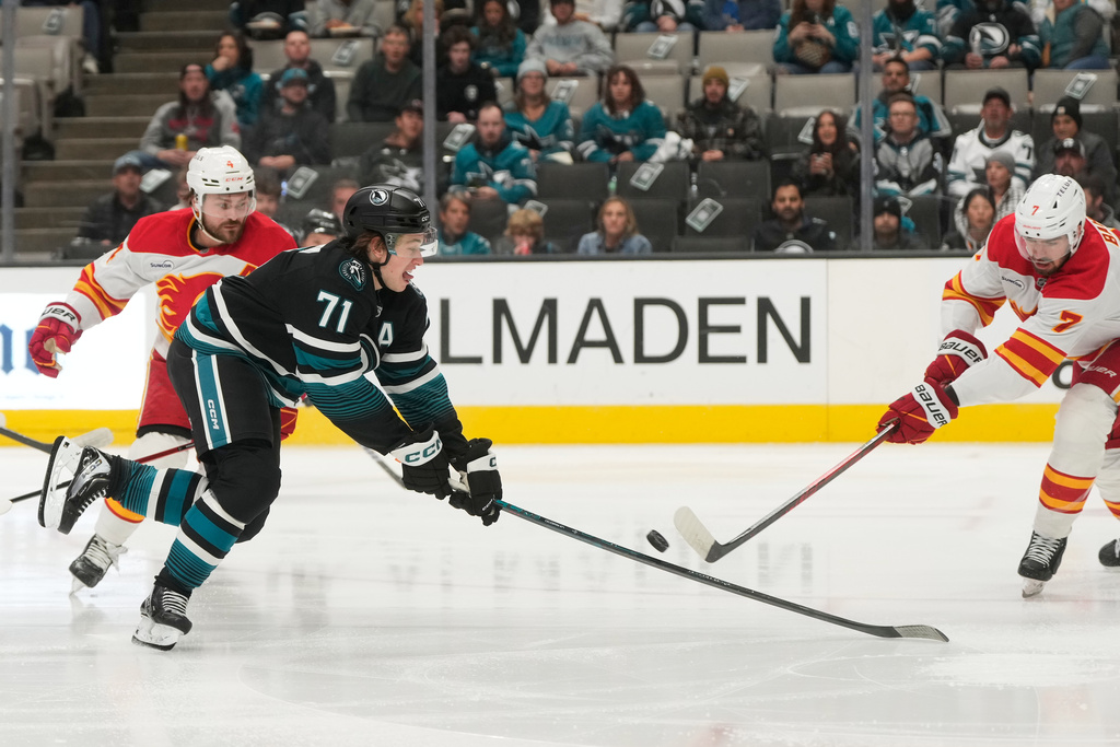 San Jose Sharks center Macklin Celebrini (71) reaches for the puck against Calgary Flames defenseman Rasmus Andersson, left, and defenseman Kevin Bahl (7) during the first period of an NHL hockey game in San Jose, Calif., Tuesday, Dec. 16, 2025. (AP Photo/Jeff Chiu)