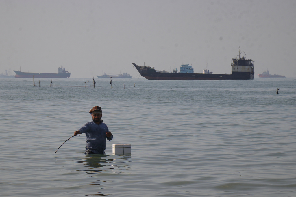 A man stands in the water, appearing to fish, as bulk carriers, cargo ships, and service vessels line the horizon in the Strait of Hormuz off Bandar Abbas, Iran, Monday, April 27, 2026.(Razieh Poudat/ISNA via AP)