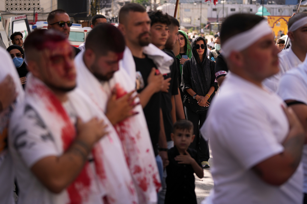 A woman watches as Lebanese Shiites beat their chests and bleed from self-inflicted ceremonial head wounds during Ashoura, when Shiites commemorate the martyrdom of Imam Hussein, the grandson of the Prophet Muhammad, amid buildings destroyed by Israeli strikes in Nabatiyeh, southern Lebanon, Friday, June 26, 2026. (AP Photo/Hassan Ammar)