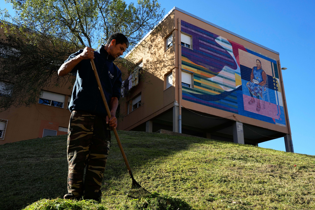 A worker rakes cut grass next to a mural by artist Carla Santiago, inspired by the United Nations Sustainable Development Goal 8, Decent Work and Economic Growth, at the housing project Zambujal in Amadora, on the outskirts of the Portuguese capital Lisbon, Thursday, Nov. 27, 2025. (AP Photo/Armando Franca)