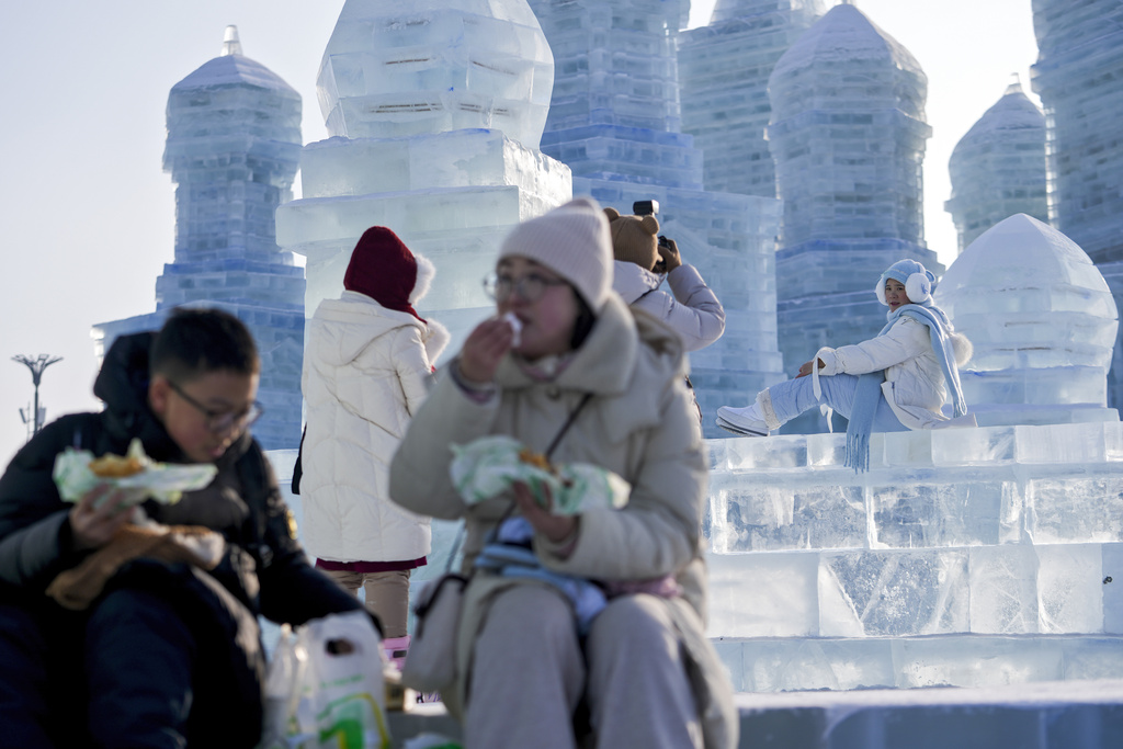Visitors eat foods as a woman poses for a photo with the ice structures at the Harbin Ice and Snow World in Harbin, China's Heilongjiang province on Monday, Jan. 6, 2025. (AP Photo/Andy Wong)
