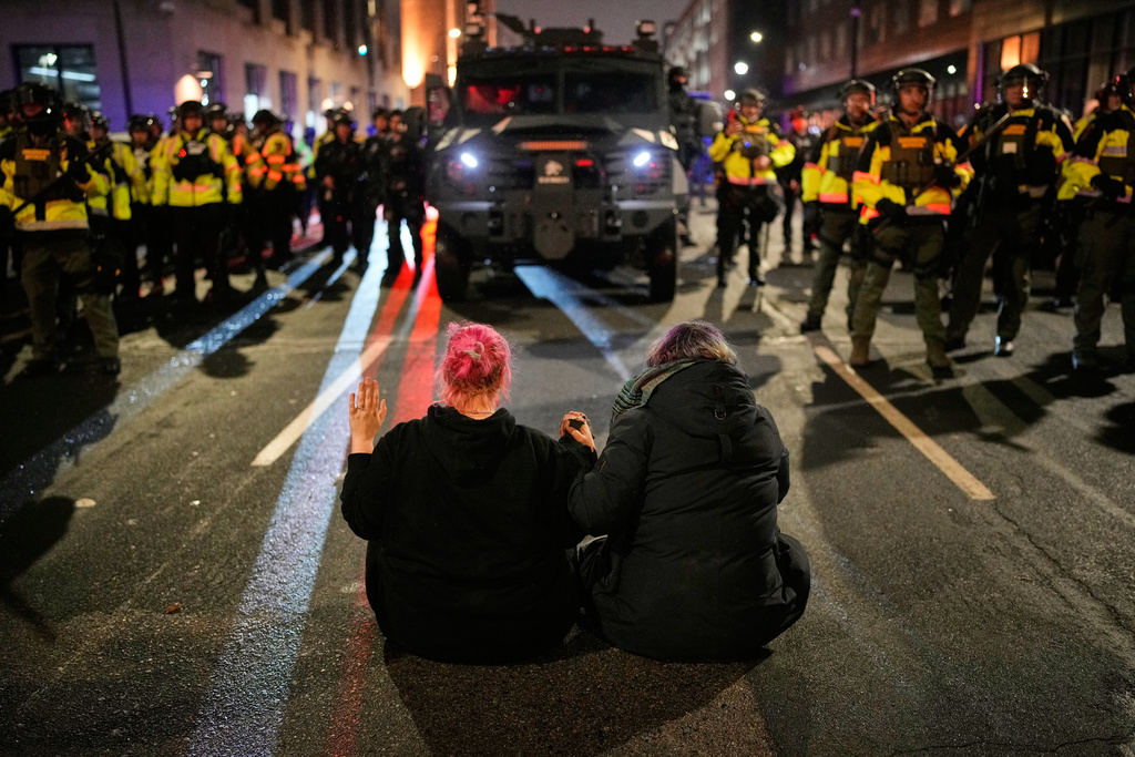Two people sit in the street holding hands in front of Minnesota State Patrol during a protest and noise demonstration calling for an end to federal immigration enforcement operations in the city, Friday, Jan. 9, 2026, in Minneapolis. (AP Photo/John Locher)