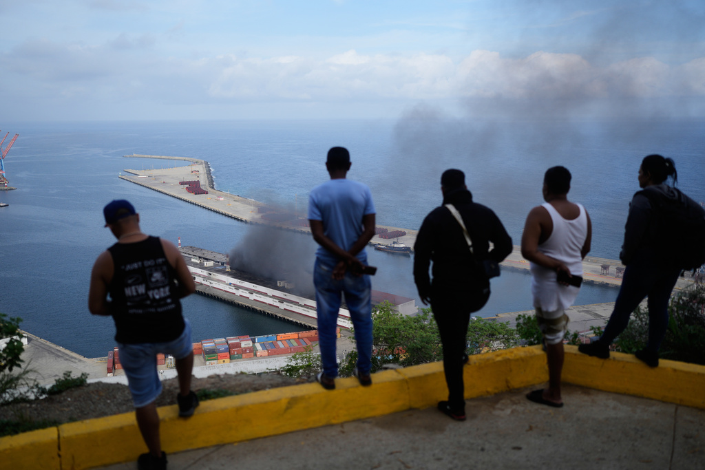 Men watch smoke rising from a dock after explosions were heard at La Guaira port, Venezuela, Saturday, Jan. 3, 2026. (AP Photo/Matias Delacroix)