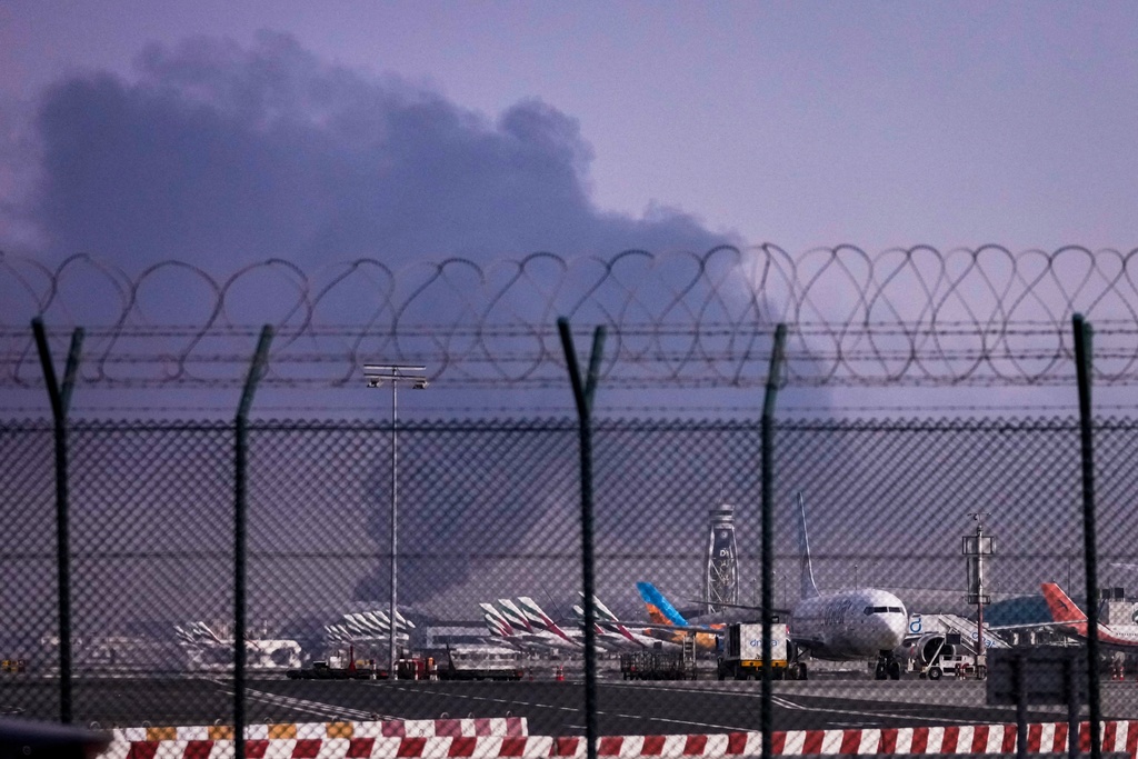 Planes are parked at Dubai International Airport as smoke rises in the background after a drone struck a fuel tank early morning, forcing the temporary suspension of flights, in Dubai, United Arab Emirates, Monday, March 16, 2026. (AP Photo)