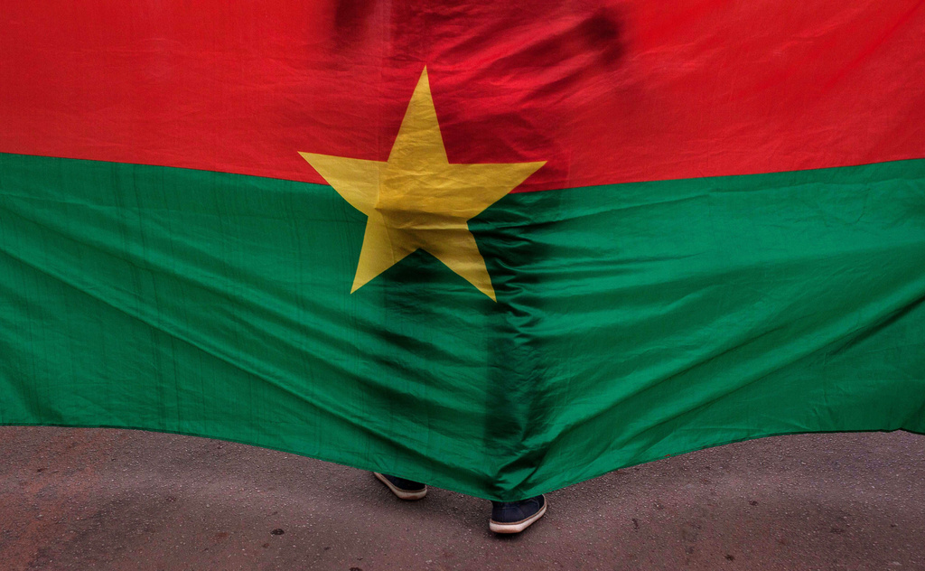 FILE - The Burkina Faso nationals flag with a man standing in front of it is seen during a memorial ceremony in Ouagadougou, Burkina Faso, Jan. 23, 2016. (AP Photo/Theo Renaut, File)