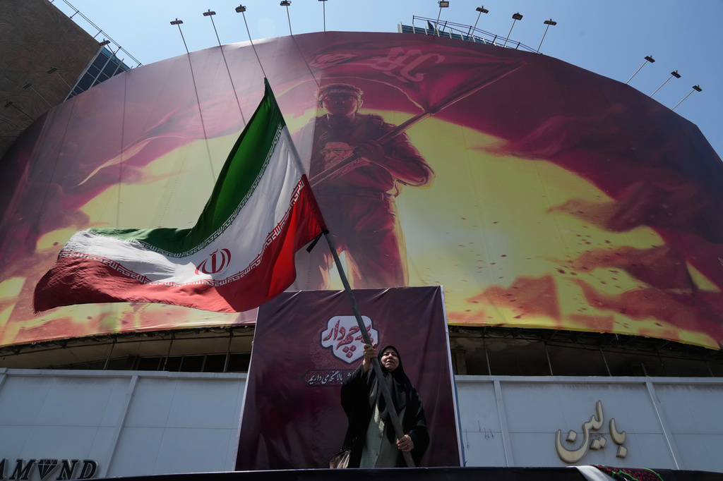 A woman waves an Iranina flag next to a billboard showing a graphic of a man wearing a shirt showing the face of late Iranian Supreme Leader Ayatollah Ali Khamenei, who was killed during a joint U.S. and Israeli strike on Feb. 28, with words calling upon the avengers of Imam Hussein, one of Shiite Islam's most beloved historical saints, in a square in downtown Tehran, Iran, Saturday, Aug. 1, 2026. (AP Photo/Vahid Salemi) CORRECTS: Amends wording for clarity.