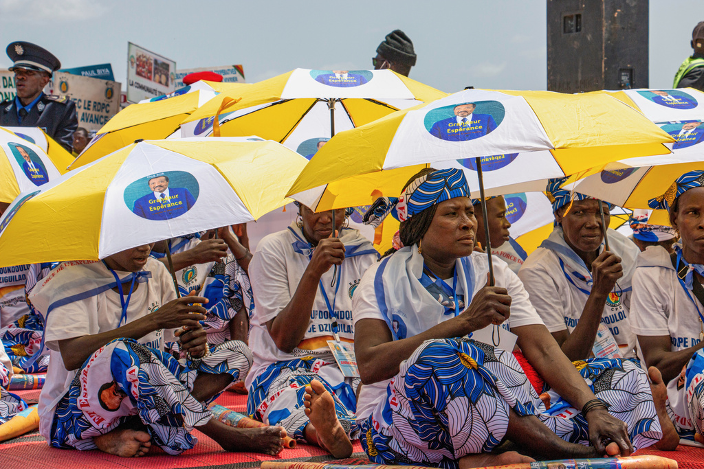 Women dressed in matching patterned outfits sit under umbrellas bearing the image of President Paul Biya during a political rally at Lamido Yaya Dairou Stadium in Maroua, Cameroon, Tuesday, Oct. 7, 2025. (AP Photo/Welba Yamo Pascal)