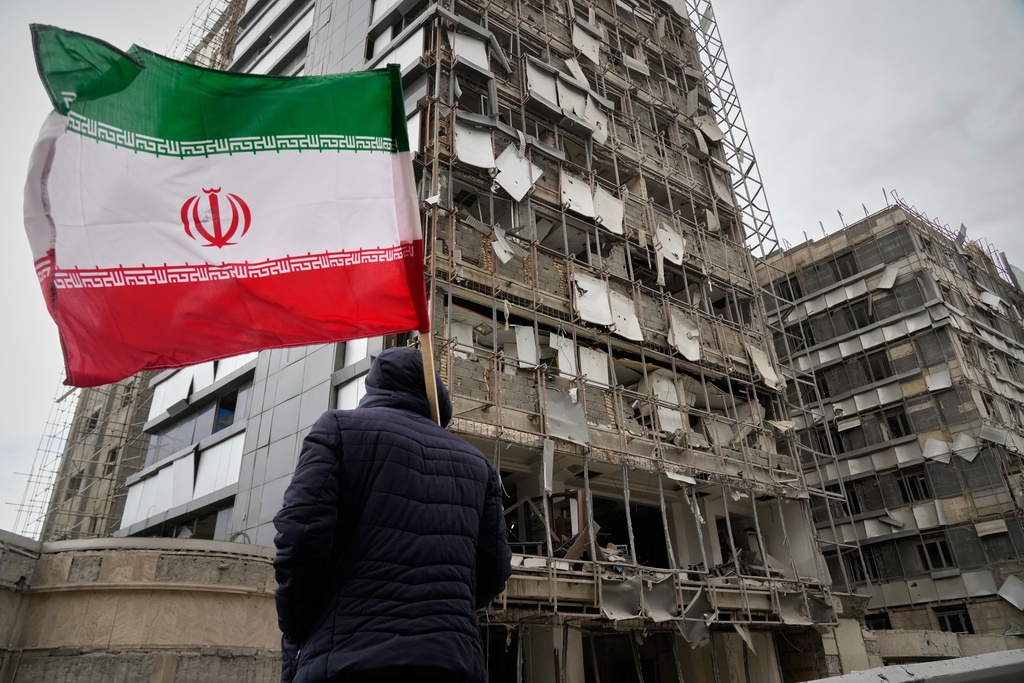 A man holds an Iranian flag as he looks at the damaged façade of Gandhi Hospital, which was hit Sunday when a strike also struck a state TV communications tower and nearby buildings across the street during the ongoing joint U.S.–Israeli military campaign in Tehran, Iran, Monday, March 2, 2026. (AP Photo/Vahid Salemi)