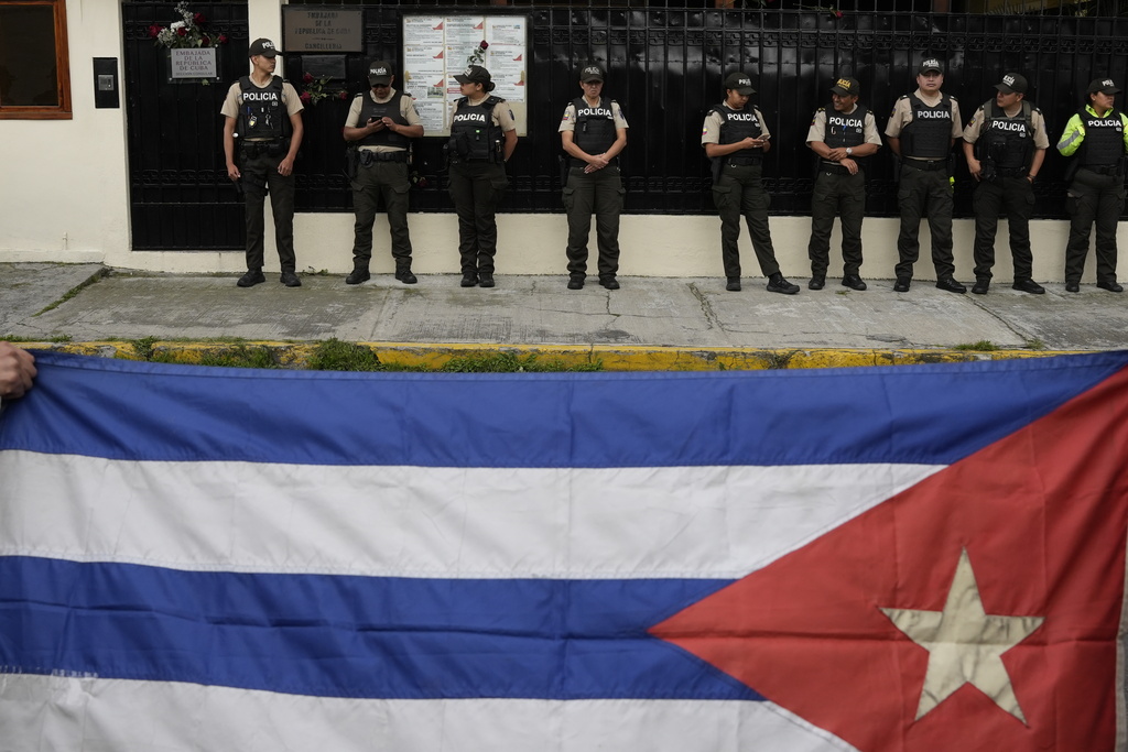 People hold up a Cuban flag as police stand guard outside the Cuban Embassy in Quito, Ecuador, Thursday, March 5, 2026. (AP Photo/Carlos Noriega)