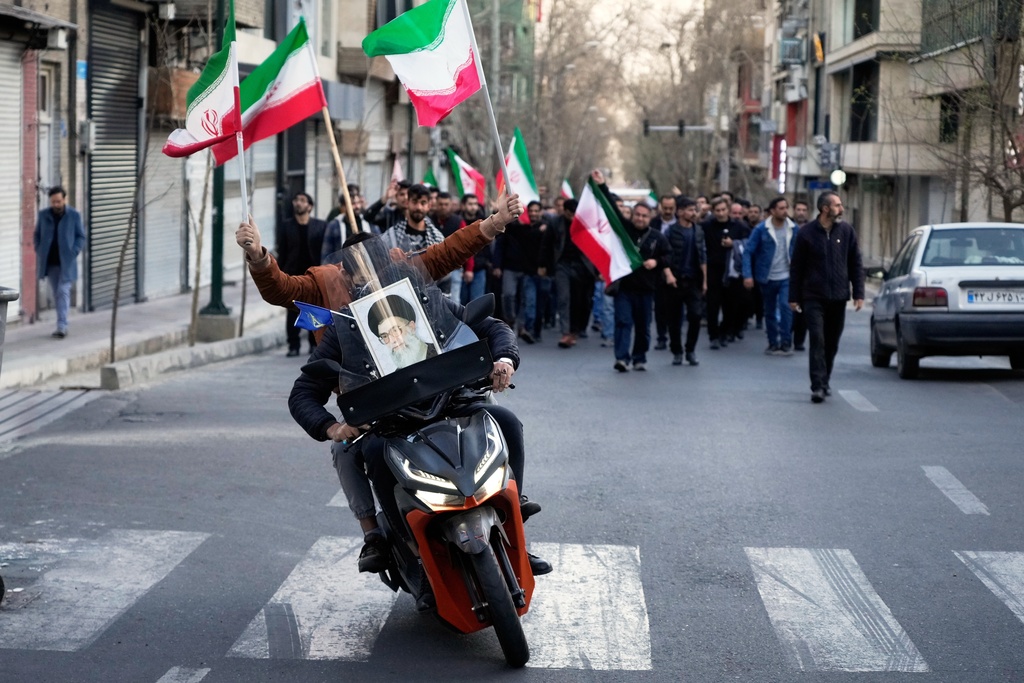 Two men on a motorcycle decorated with a picture of the late Iranian Supreme Leader Ayatollah Ali Khamenei ride ahead a group of government supporters near Khamenei's residency in Tehran, Iran, Sunday, March 1, 2026, in the aftermath of his confirmed death in U.S. and Israeli strikes. (AP Photo/Vahid Salemi)