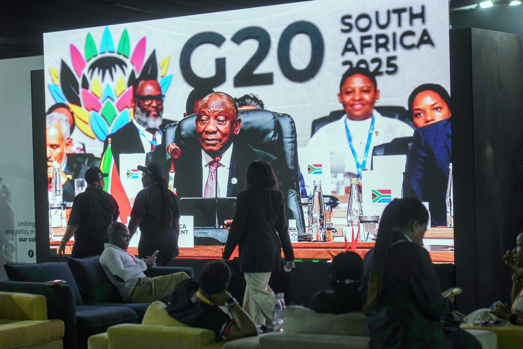 People walk by a large screen TV where South African President Cyril Ramaphosa holds a wooden gavel as he officially closes the G20 leaders' summit, in Johannesburg, South Africa, Sunday, Nov. 23, 2025. (AP Photo/Jerome Delay)