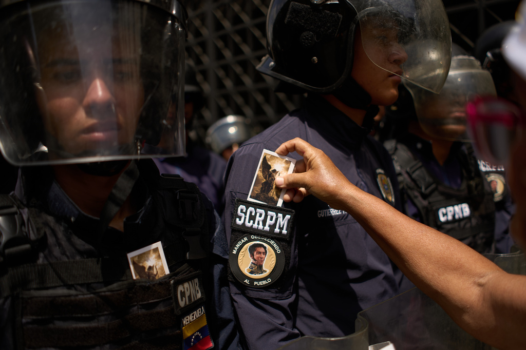 A woman places a religious card on the sleeve of a Bolivarian National Police officer, whose sleeve has a quote and photo of Independence hero Simon Bolivar, as officers stand guard by a protest of retirees asking for better health and pension conditions in Caracas, Venezuela, Friday, May 29, 2026. (AP Photo/Ariana Cubillos)