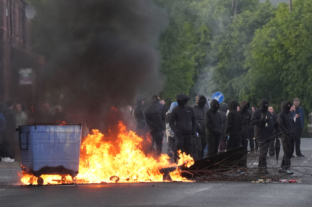 Masked protesters stand by burning trash containers on Ligoniel Road in Belfast, Northern Ireland, on Tuesday, June 9, 2026, after the arrest of a Sudanese man accused of stabbing a man in the northern part of the city. (PA via AP)