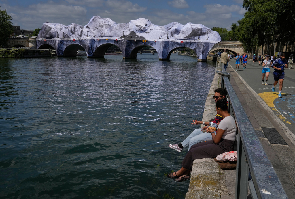 People sit along the Seine river next to "The Pont Neuf Cave," an inflated art installation by French street artist JR, on Paris' oldest bridge, the Pont Neuf, Thursday, May 21, 2026, which will be open to the public from June 6-28. (AP Photo/Michel Euler)