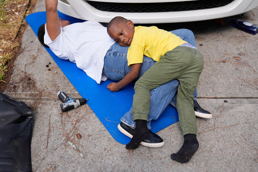 Sechita McNair, works on her broken van as her son Malachi McNair-Nesbitt lays on her in the driveway of their rental home on June 6, 2025, in Jonesboro, Ga. (AP Photo/Brynn Anderson)