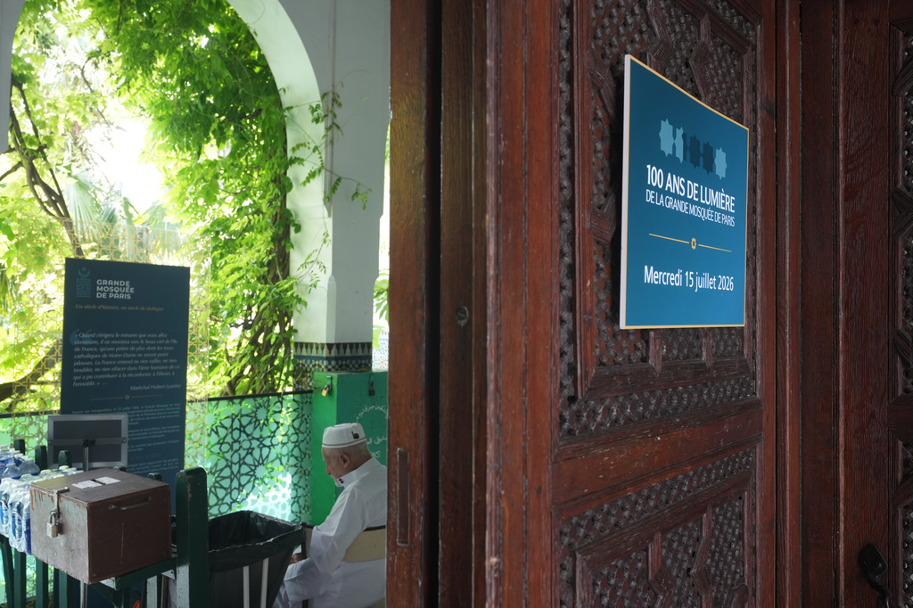 A man sits at the entrance of Paris' Grand Mosque where a blacard reads "100 Years of Light" marking its centenary, Friday, July 17, 2026. (AP Photo/Michel Euler)