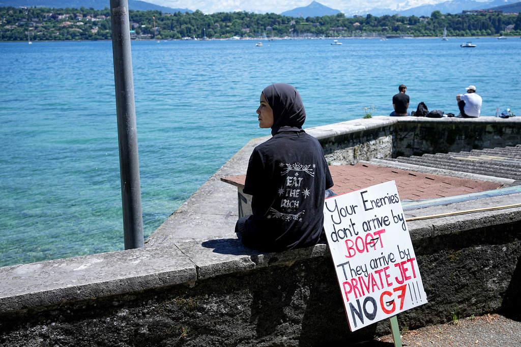 A woman sits with a sign during a demonstration against the G7 in Geneva, Switzerland, Sunday, June 14, 2026, ahead of the G7 summit scheduled to take place June 15-17. (AP Photo/Laurent Cipriani)