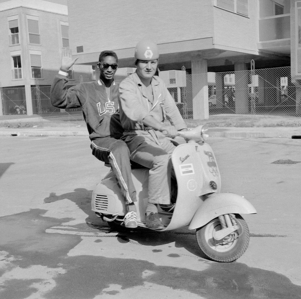 FILE - U.S. high jumper John Thomas, of Cambridge, Mass., waves from a Vespa scooter with an unidentified official Olympic driver inside the Olympic Village, in Rome, Aug. 27, 1960. (AP Photo/Mike Stern, File)