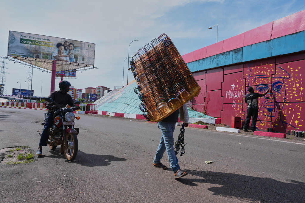 Passersby look on as graffiti artist Omar Diaw works on a mural depicting Guinea's President Mamadi Doumbouya on a street wall in Conakry, Guinea, Saturday, Sept. 20, 2025. (AP Photo/Misper Apawu)