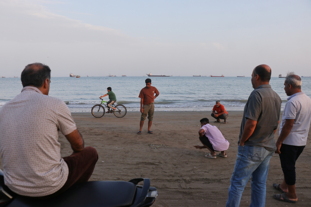 Men gather along the shore, some crouching and watching a game, as a mix of bulk carriers, cargo ships, and service vessels line the horizon in the Strait of Hormuz off Bandar Abbas, Iran, Monday, April 27, 2026.(Razieh Poudat/ISNA via AP)