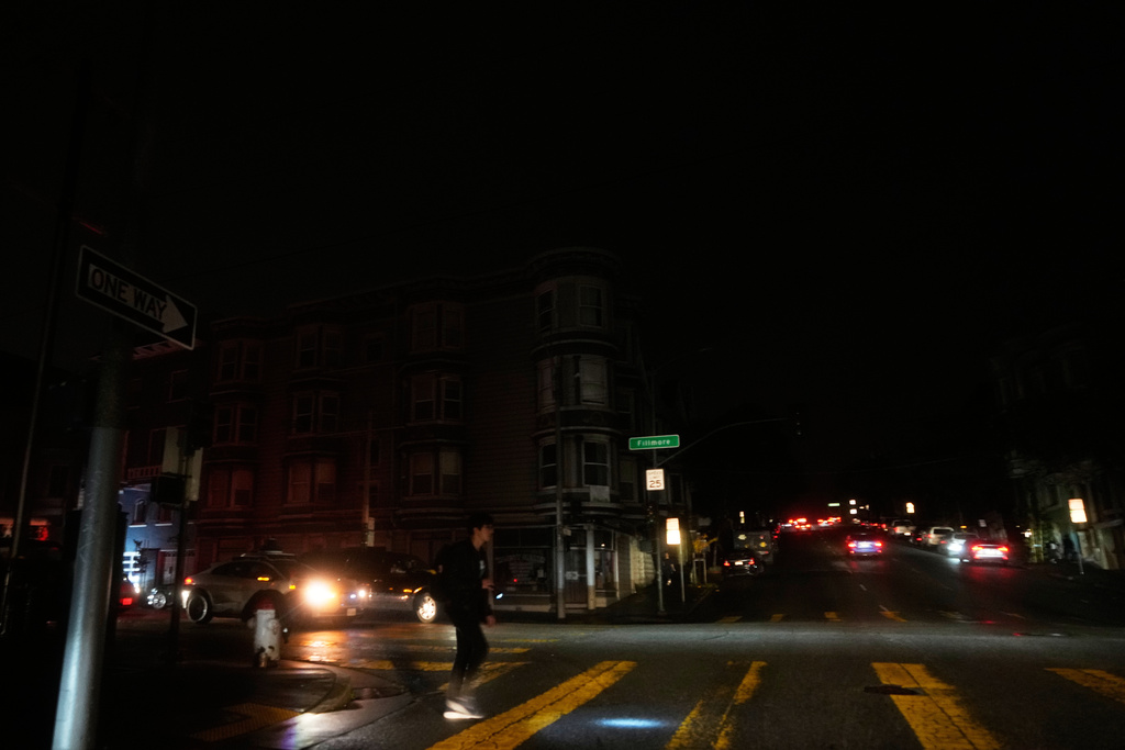 A pedestrian uses a flash light while crossing an intersection as power outages darken the streets, in San Francisco, Saturday, Dec. 20, 2025. (AP Photo/Jeff Chiu)