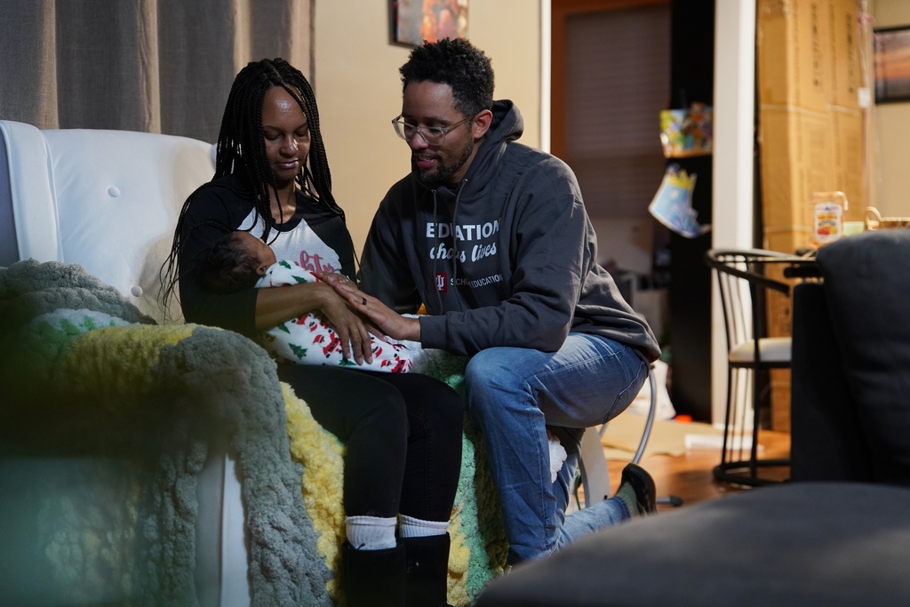 Crystal Wilmot-Burton holds their sleeping newborn daughter, Phoenix RyZen Reign Burton, as her husband, JaKobi Burton, kneels next to her at their home in Indianapolis, Dec. 12, 2025. (AP Photo/Obed Lamy)