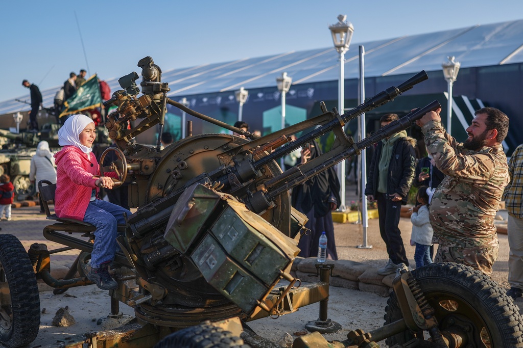 A girl sits on a machine gun as visitors tour the "Syrian Revolution Military Exhibition," which opened last week ahead of the first anniversary of the ousting of the Bashar Assad regime in Damascus, Syria, Sunday, Dec. 7, 2025. (AP Photo/Ghaith Alsayed)